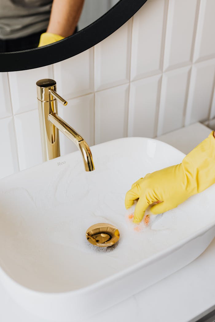 Person cleaning a bathroom sink with foam and yellow gloves, emphasizing hygiene and cleanliness.